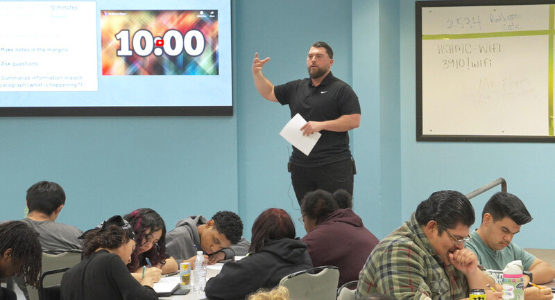 Screenshot from video of teacher standing in front of a lecture-style high school class, gesturing and talking to students