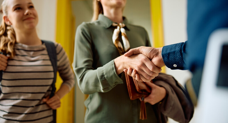 A close-up of a mother standing beside her teenage daughter as they meet and shake hands with the teacher.